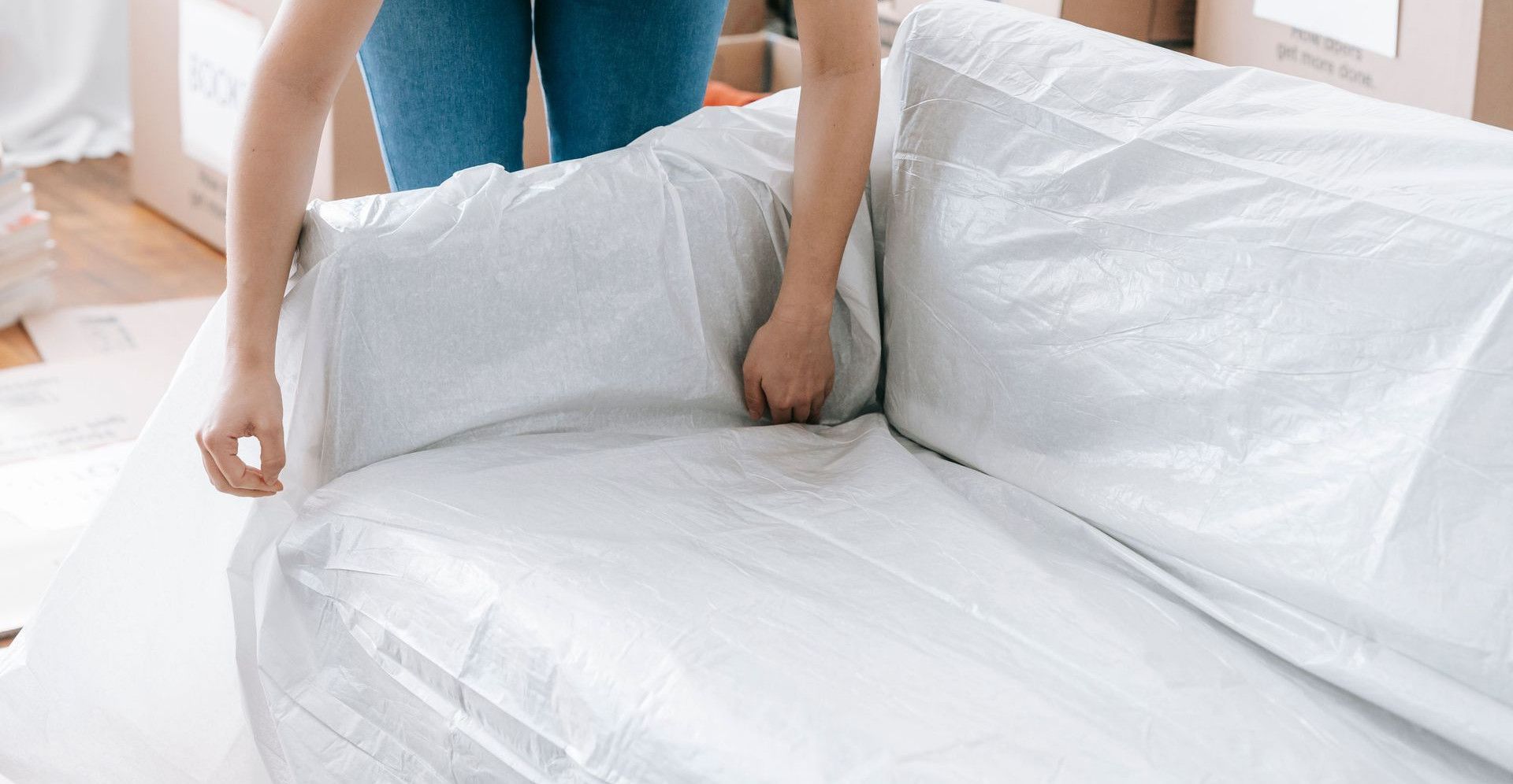A woman carefully wraps a sofa in protective material while packing boxes in a bright room.